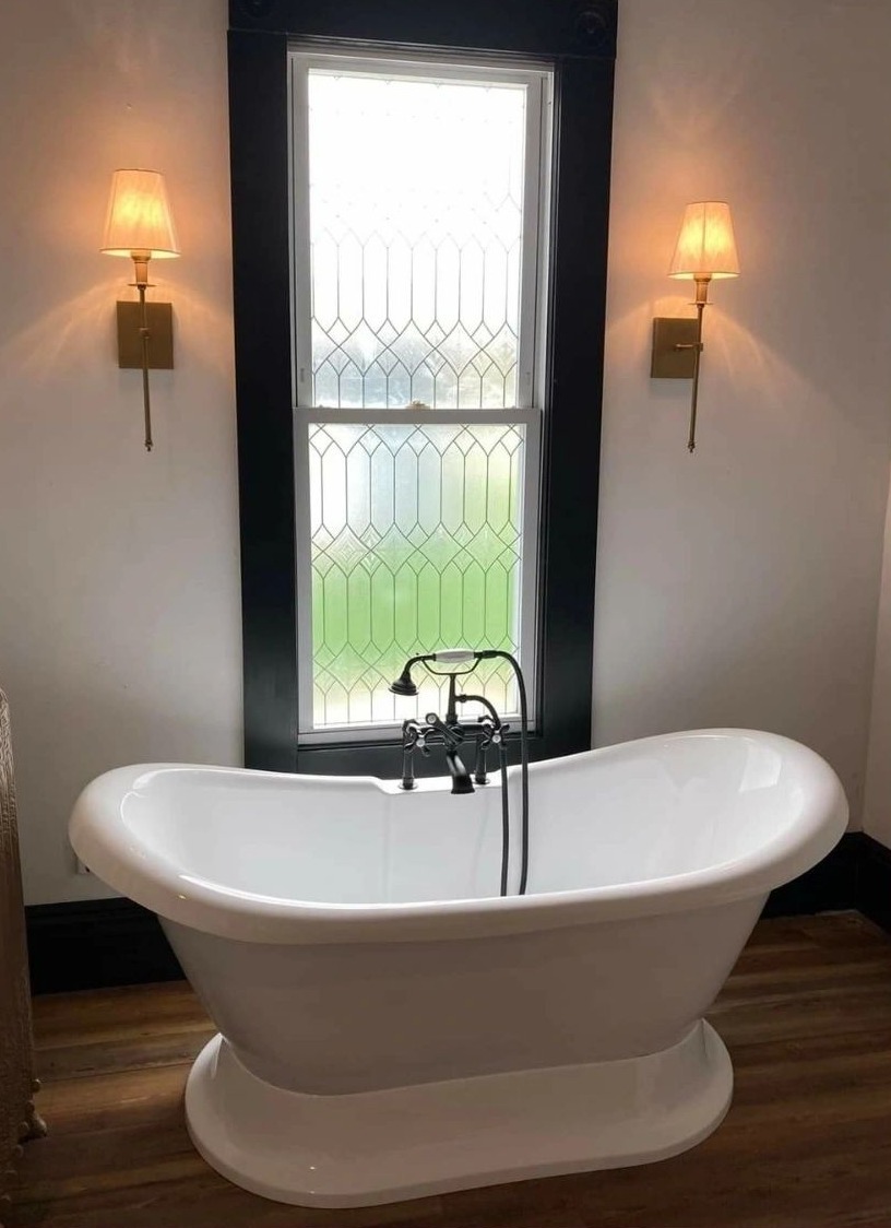 A freestanding double-slipper tub under a tall leaded-glass window, with a wall sconce either side and a tap and handshower at the rim.